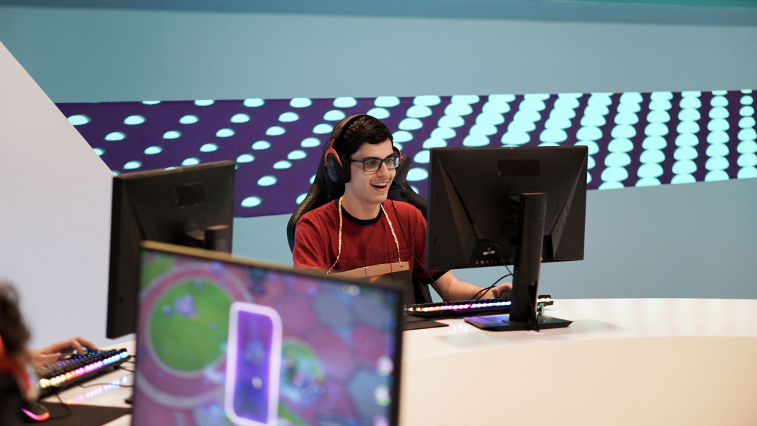 Person sitting behind his laptop in a big gaming events hall gaming with other people. He has a smile on his face.