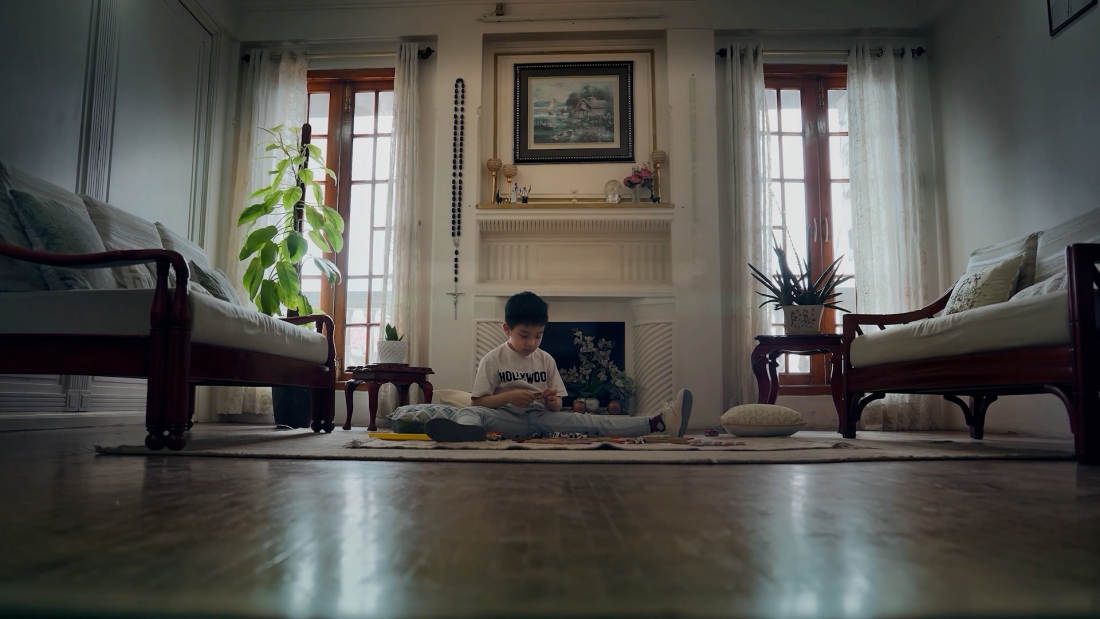 The image shows a young boy sitting on a carpeted floor in a well-lit, cozy living room, playing with toys.