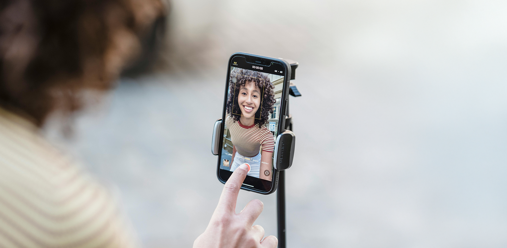 A close-up image of a smartphone mounted on a tripod, capturing a cheerful woman with curly hair wearing a striped shirt during a video recording. A hand is visible in the foreground, about to tap a button on the phone screen.