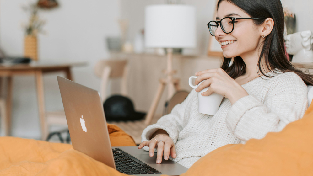 Women sitting on a couch looking at a laptop screen and she has a cup in her hand.